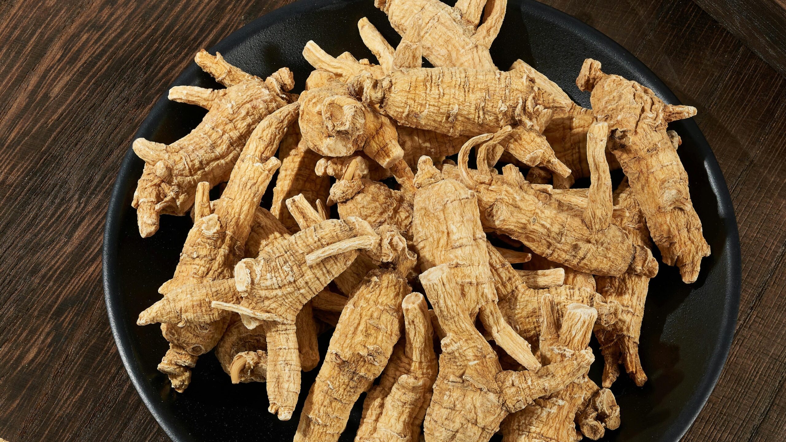 High angle view of fresh ginseng roots on a black plate on a wooden table, showcasing the natural texture and earthy tone.