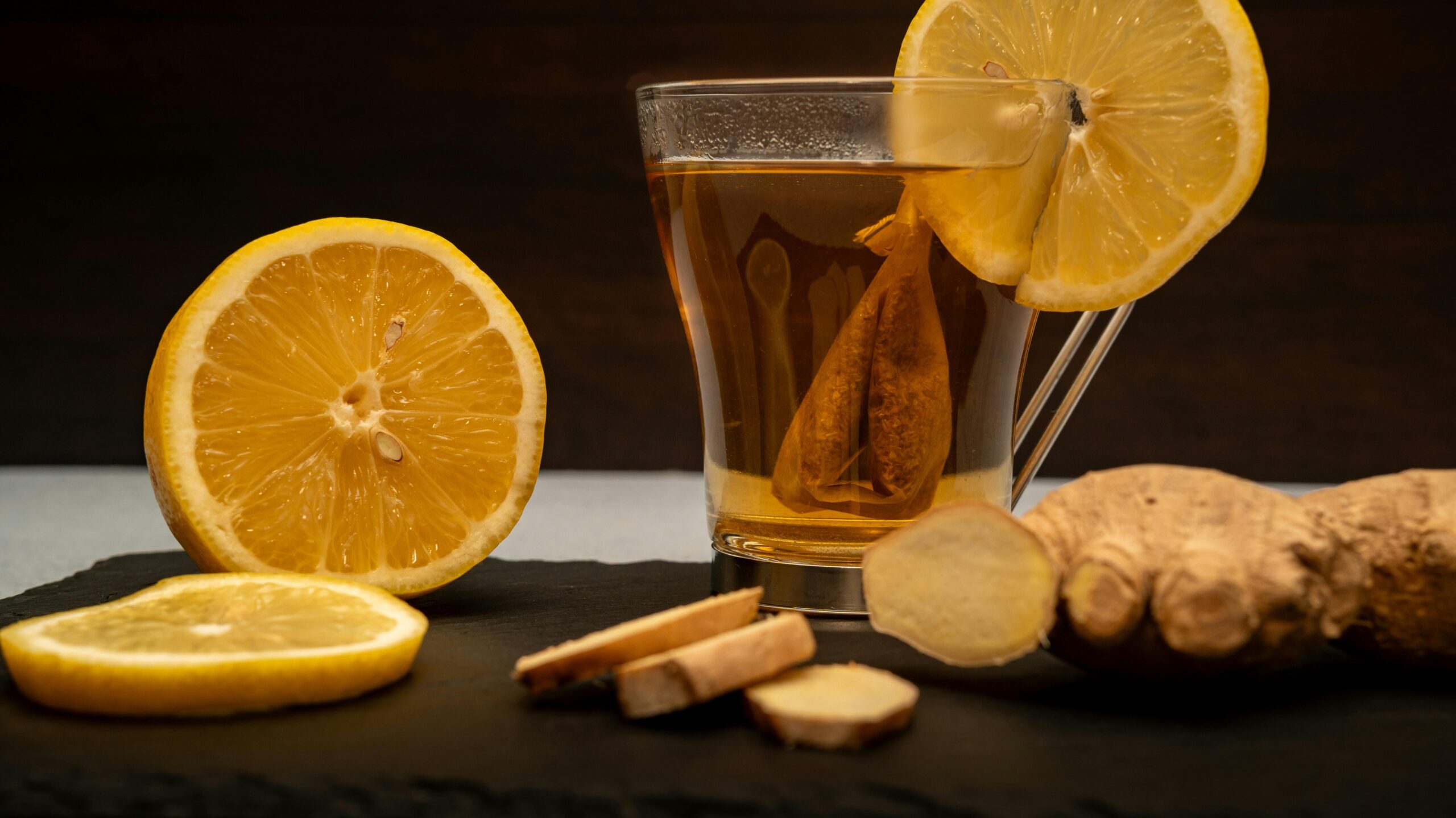A warm cup of lemon ginger tea surrounded by fresh lemon slices and ginger root on slate.