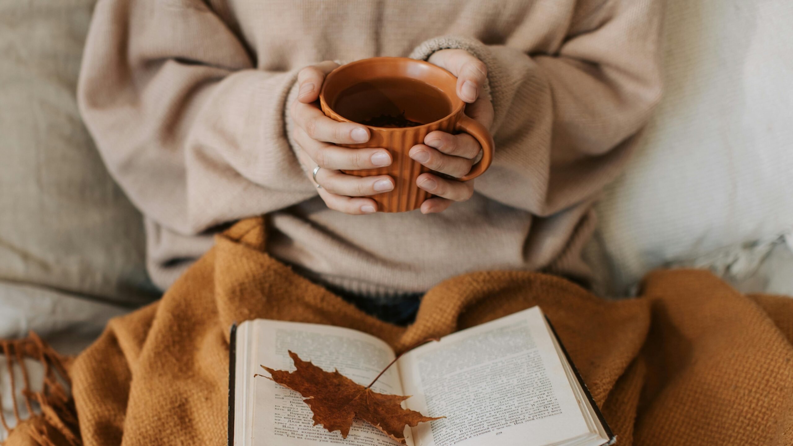 A woman enjoys a cozy autumn day, holding a tea mug over an open book with a maple leaf.