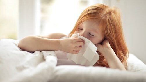 Redheaded child resting in bed, wiping nose with tissue, unwell with flu-like symptoms.